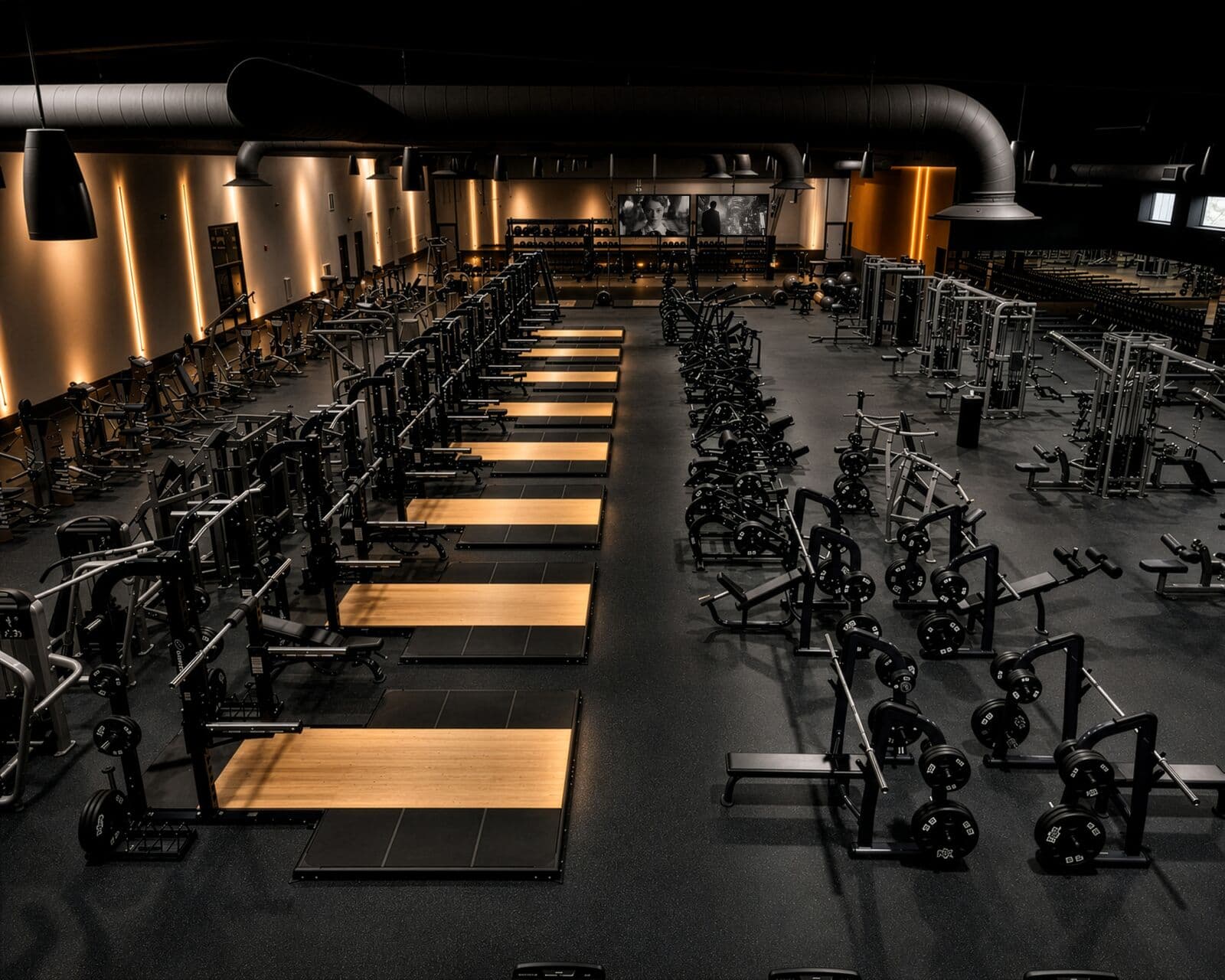 Elevated wide view of the Sanctuary Fitness strength floor, with wood lifting platforms, plate-loaded machines, and warm accent lighting along the walls.